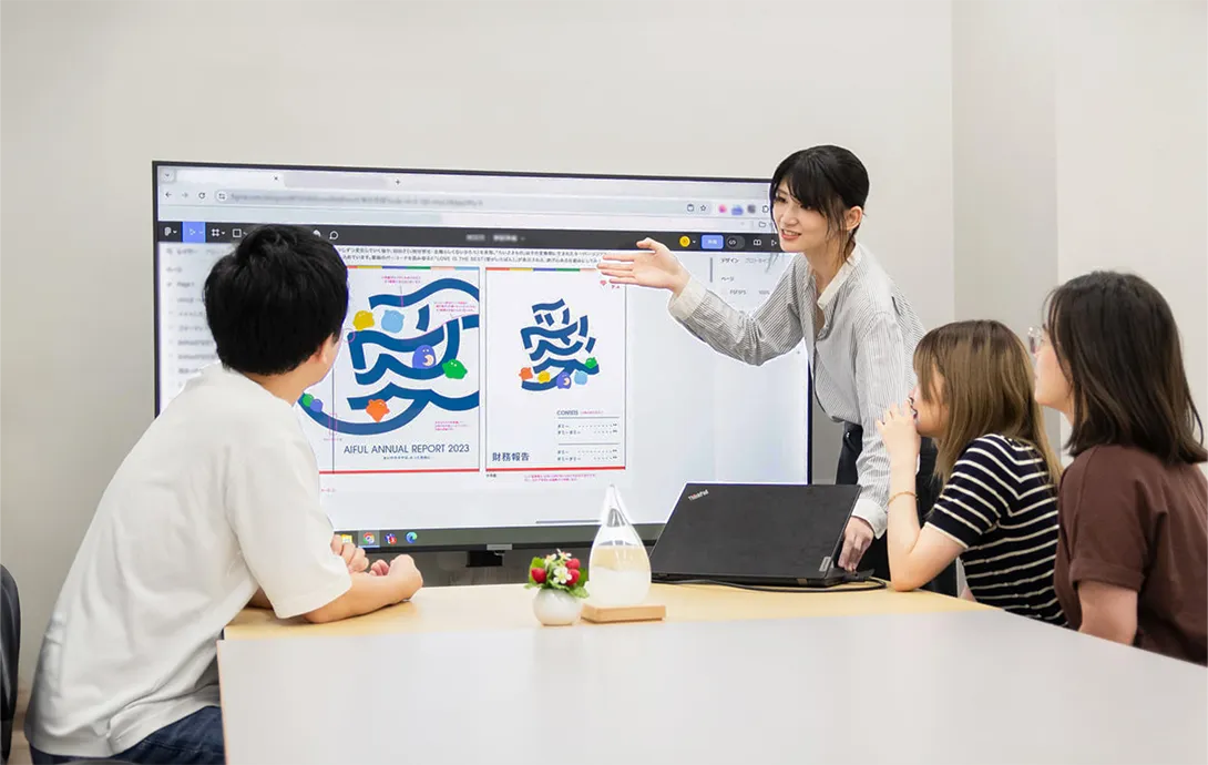 デザインデータを表示した大きなモニターを前に説明をするデザイナー社員と話を聞く3名